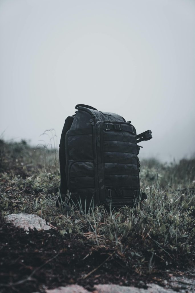 A rugged black backpack sits on grass in a misty outdoor environment, evoking a sense of adventure.