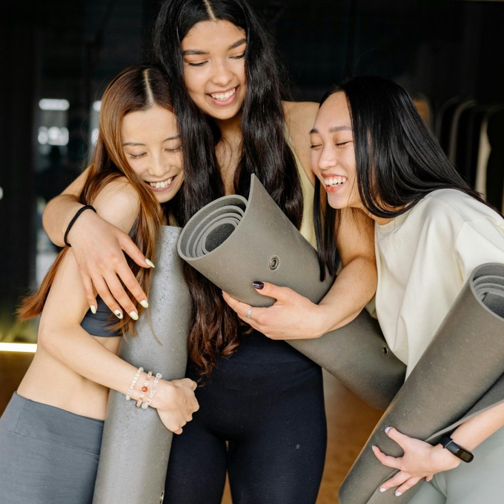 Three young women joyfully hugging and holding yoga mats, symbolizing friendship and healthy living.