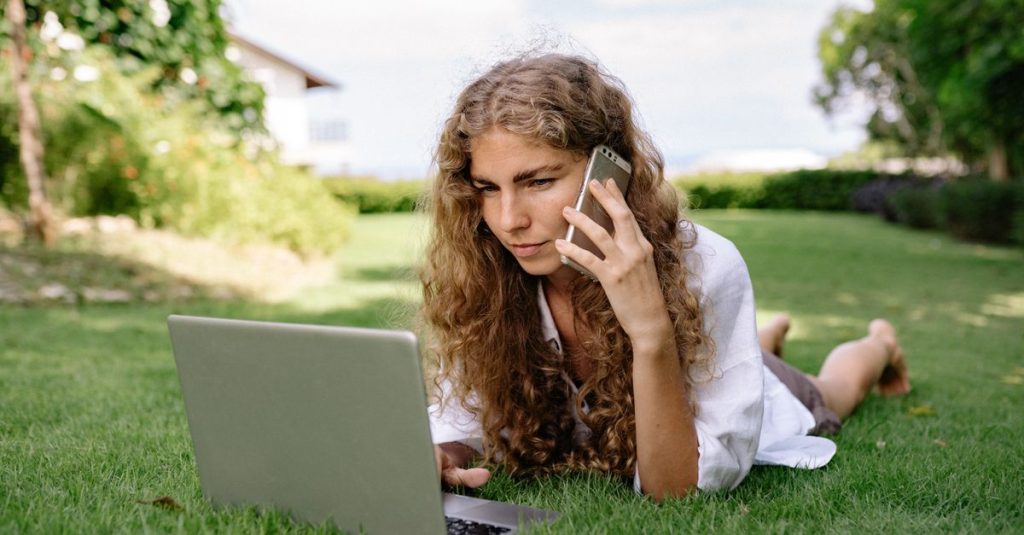 A young woman multitasks on a laptop and smartphone while lying on green grass outdoors.