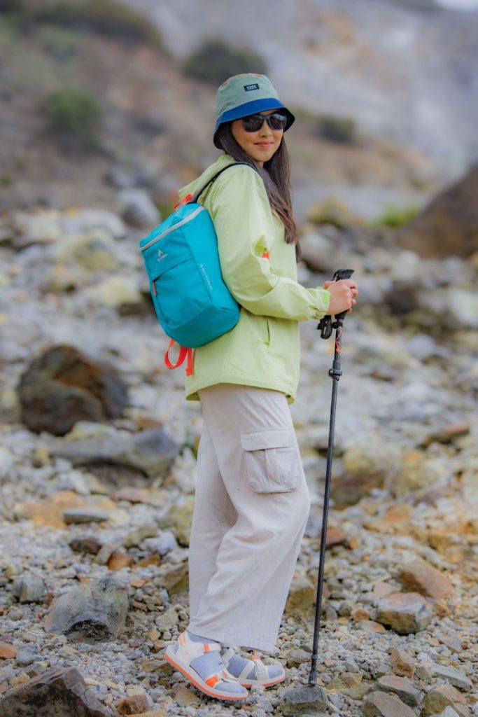 Woman hiking outdoors in rocky terrain with a teal backpack, bright clothing, and walking stick.