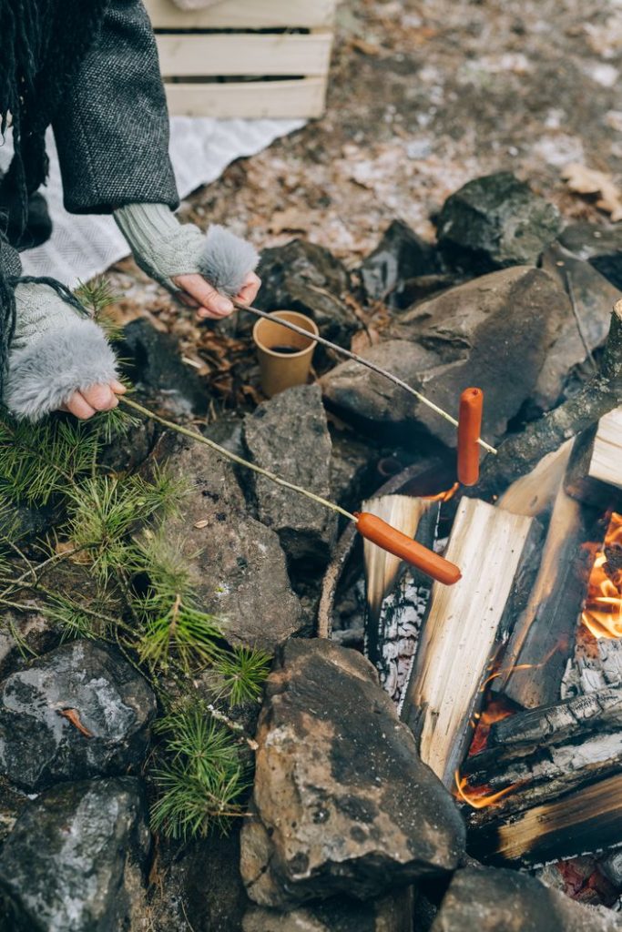 A person roasts sausages over a campfire outdoors, surrounded by rocks.