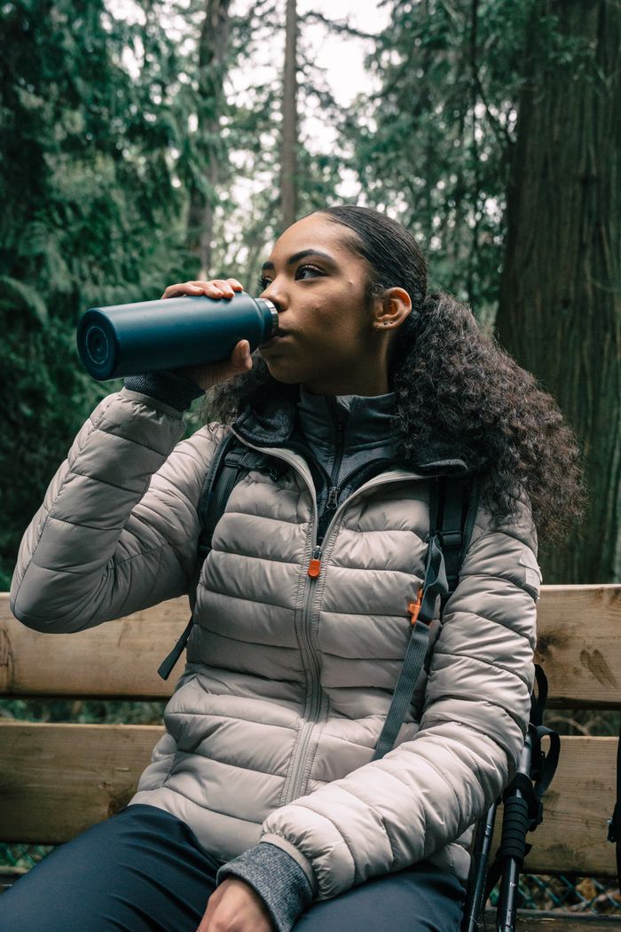 A woman sitting on a bench in a forest drinking from a tumbler, dressed in hiking gear.