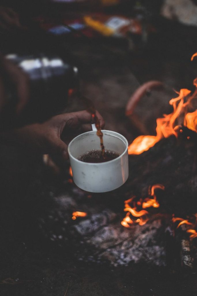 A rustic scene of coffee being poured over a campfire, evoking warmth and adventure.